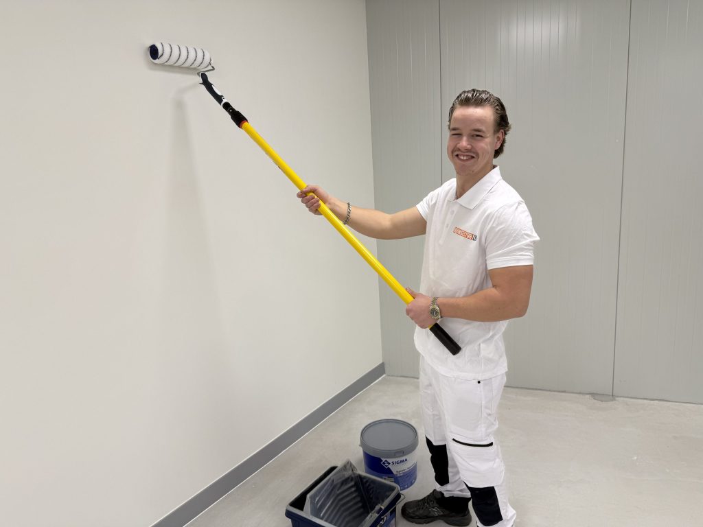 Lachende schilder in witte werkkleding met een gele verlengsteel en een emmer Sigma-verf aan zijn voeten