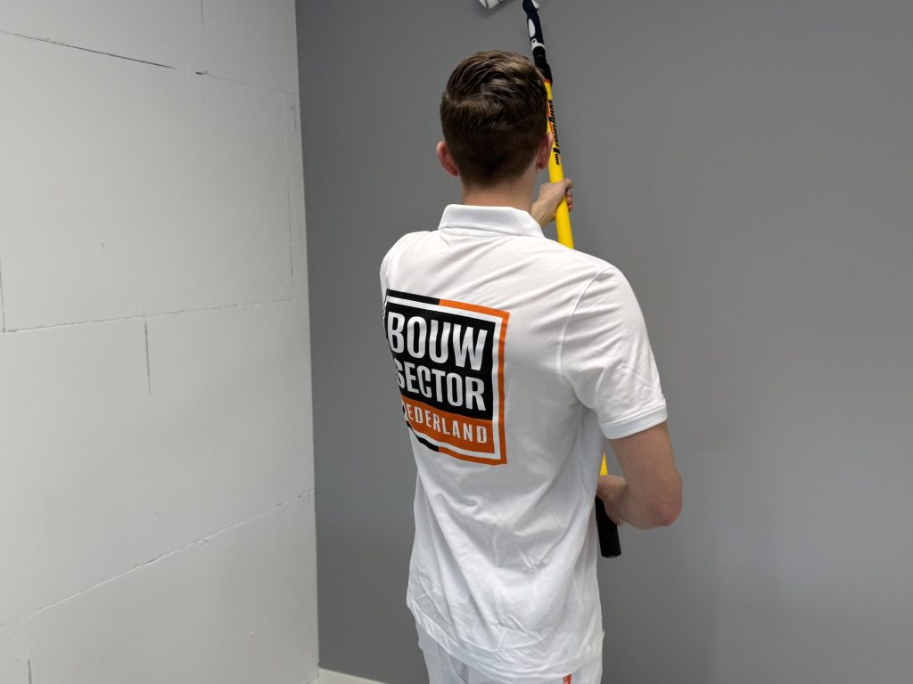 Schilder rolt een grijze muur met een lange roller, naast een nog kale wand van blokken