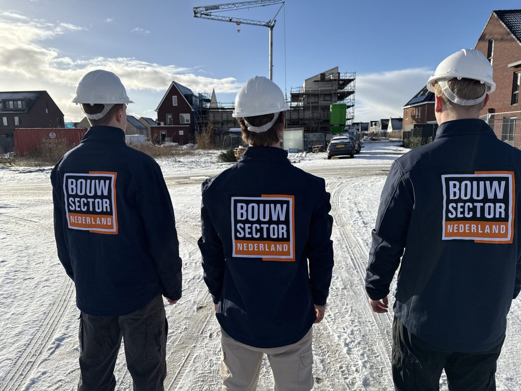 Drie vakmensen van Bouwsector Nederland met witte helm en logo op de jas staan op een besneeuwde bouwweg tussen nieuwe huizen